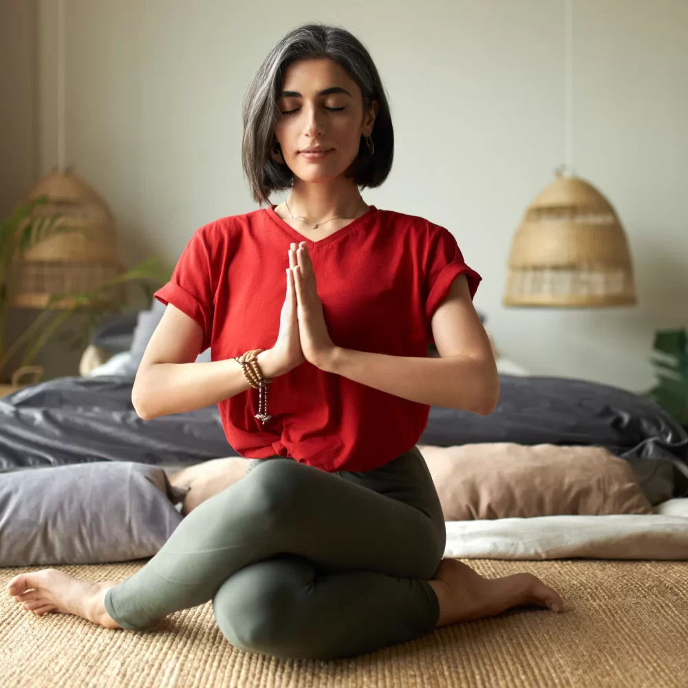 Fashionable young female with grayish hair sitting in gomukhasana or cow pose while practicing hatha yoga in bedroom after awakening, keeping eyes closed, pressing hands together in namaste